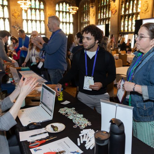 Color photo of a sponsor representative engaged in conversation with two marketers at BrandSmart 2025; other attendees are seen in the background, conversing in the Cathedral Room at University Center